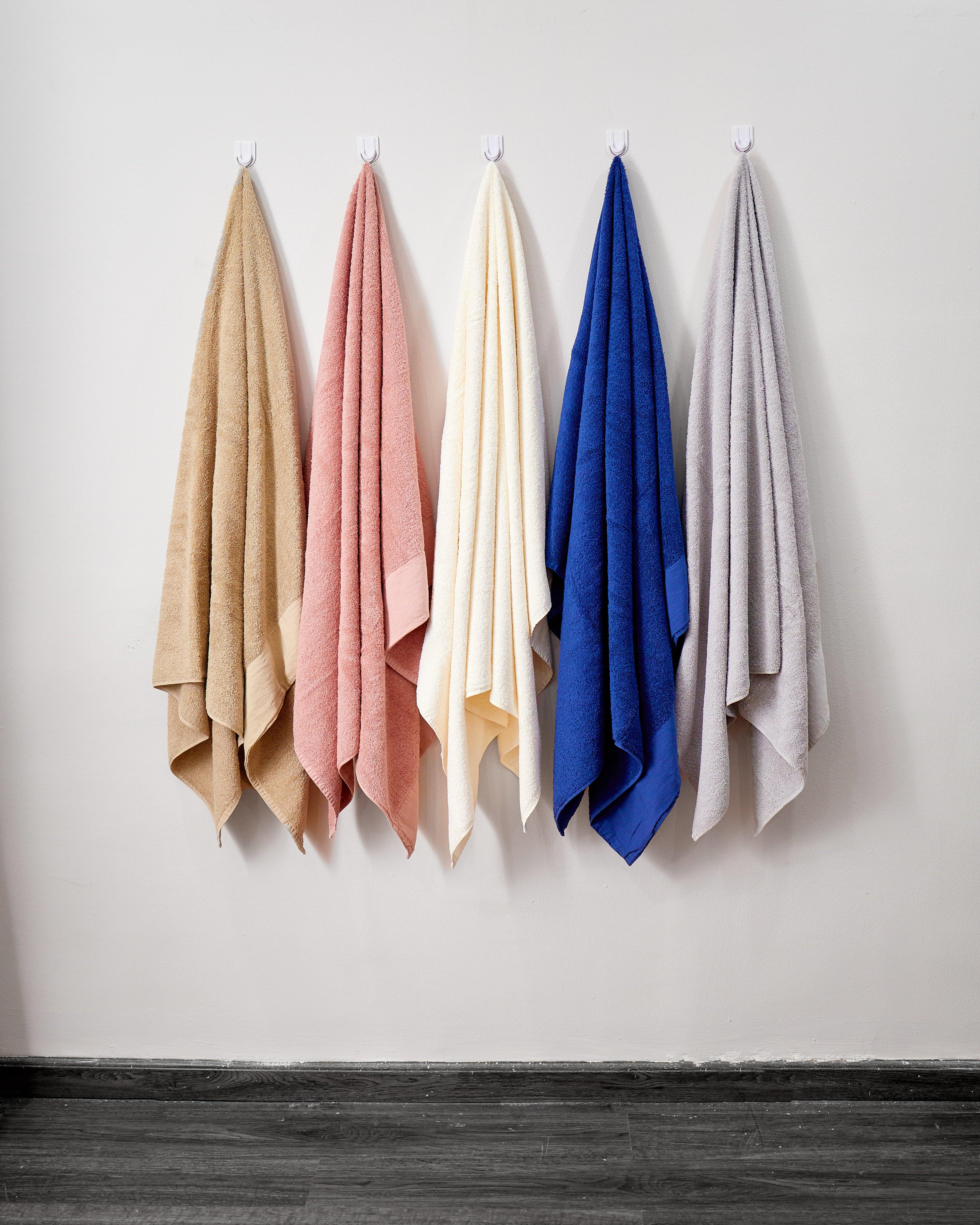 Five Drench collection towels in beige, pink, cream, blue, and gray hanging on hooks against a white wall