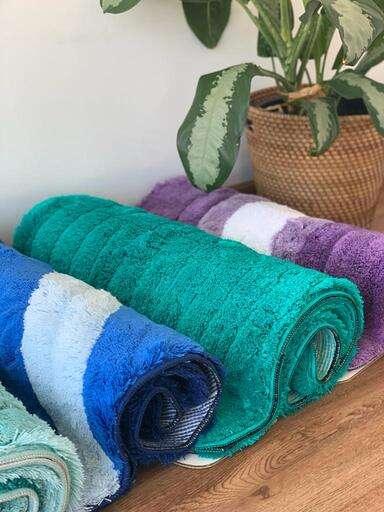 Rolled Lory Single Bath Rugs in various colors with plush texture and non-slip backing next to a wicker plant pot.